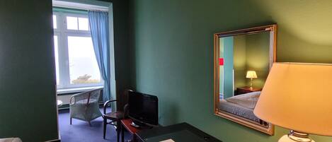 Une chambre d’hôtel comprenant un lit, un bureau avec une chaise, une lampe, un miroir et une fenêtre avec des rideaux.