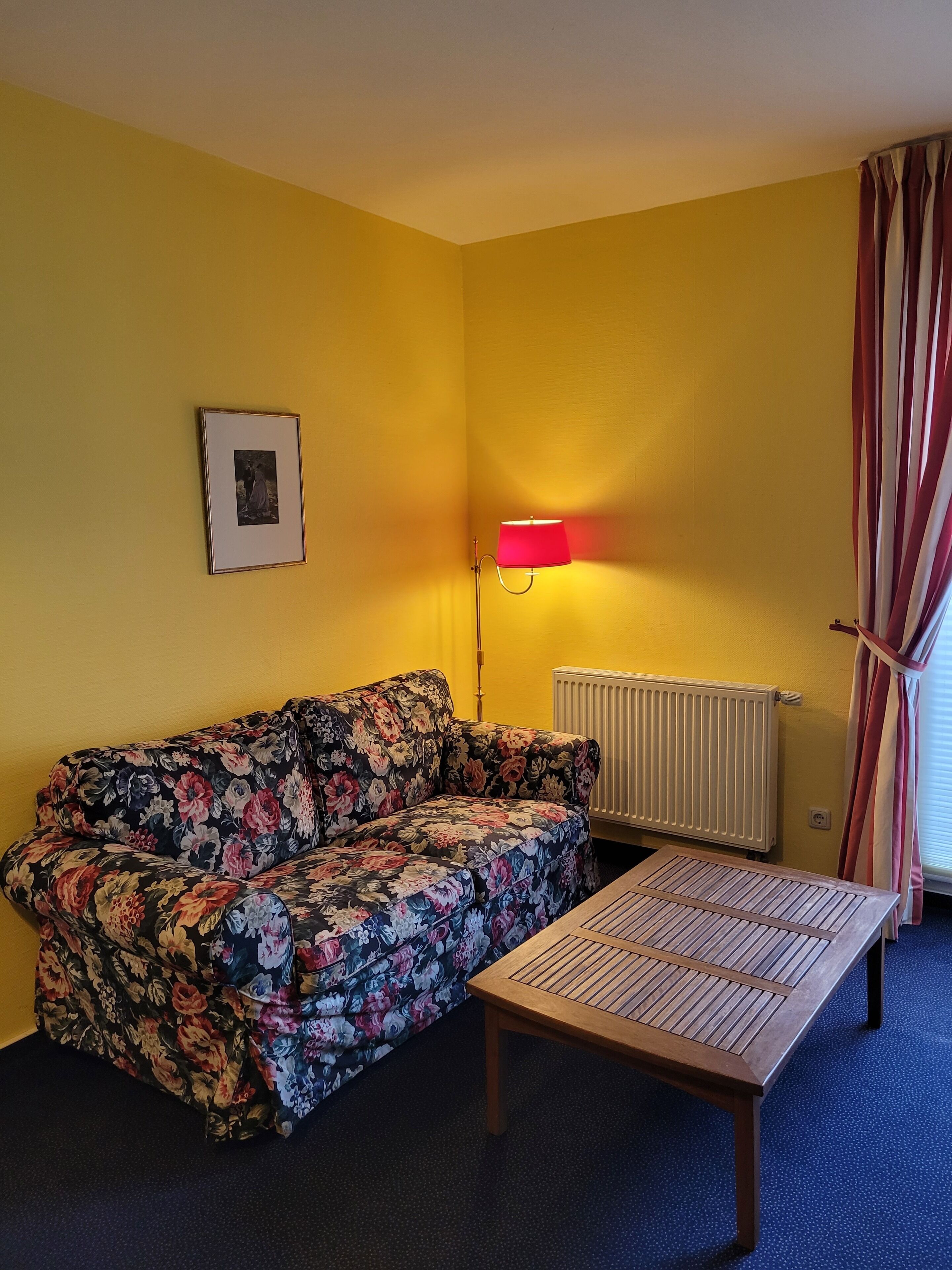 Ein Zimmer mit einem blumigen Sofa, einem Holztisch, einem Heizkörper und einer roten Lampe.