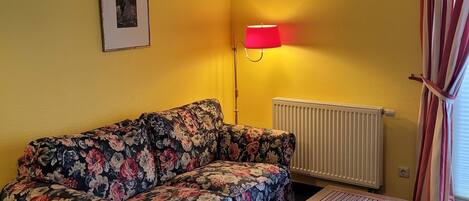 A room with a floral sofa, a wooden table, a radiator, and a red lamp.