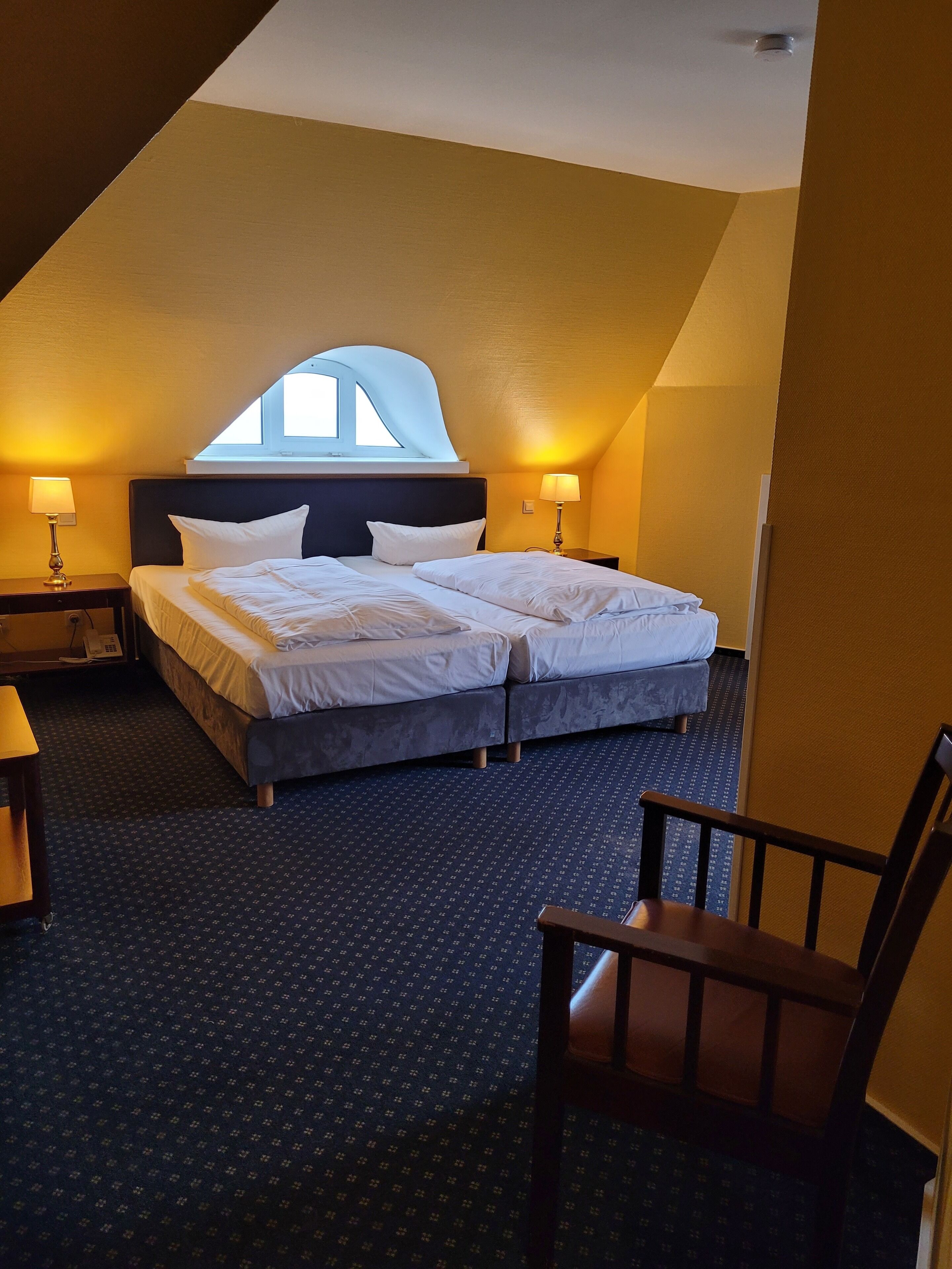 Hotellihuone, jossa on suuri sänky, kaksi yöpöytää valaisimineen, ikkuna ja kuviollinen matto.
