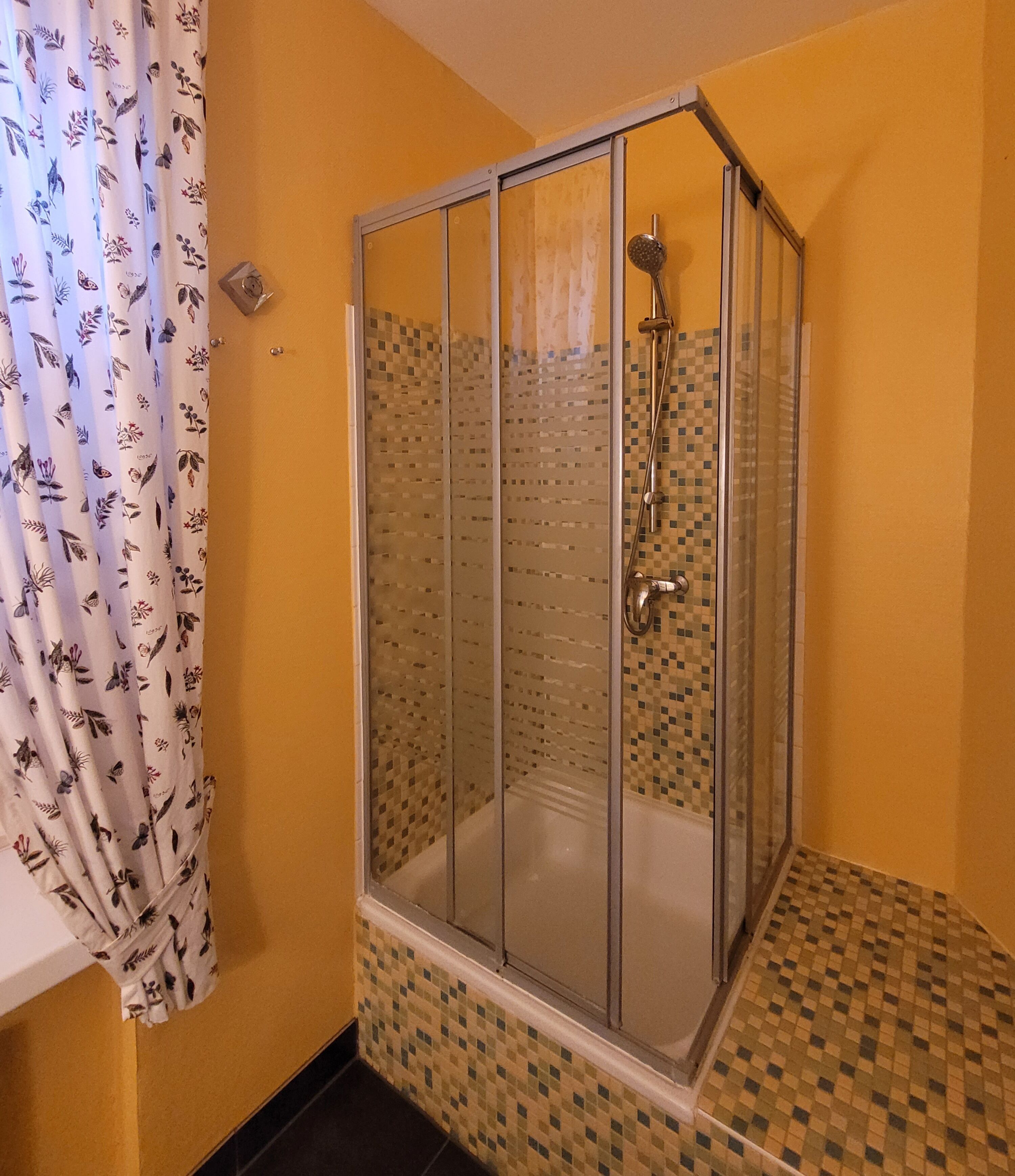 A glass-enclosed shower with a tiled floor and walls, a white curtain with a floral pattern, and a small window.