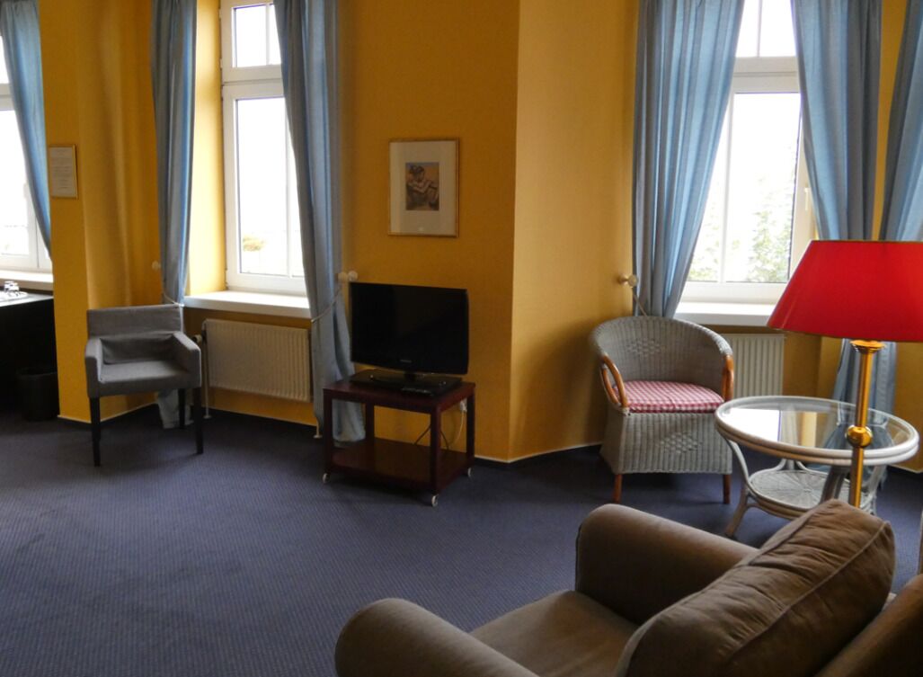 Hotellihuone, jossa on sohva, nojatuoli, televisio ja pieni pöytä, jossa on lamppu.
