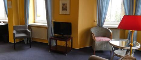 A hotel room with a sofa, armchair, TV, and a small table with a lamp.