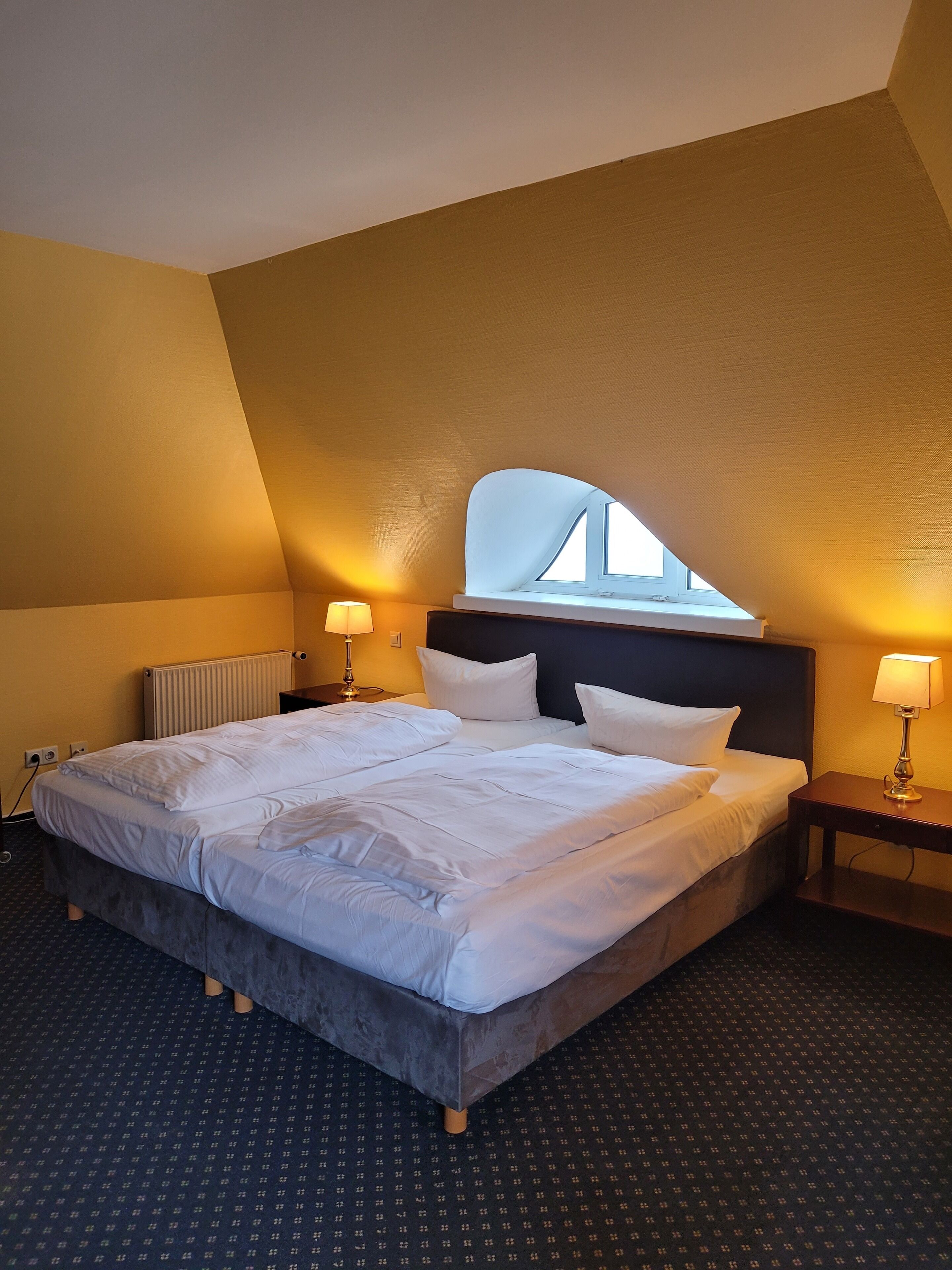 Una habitación de hotel con una cama grande, dos mesitas de noche, un ventanal con vistas y un radiador.