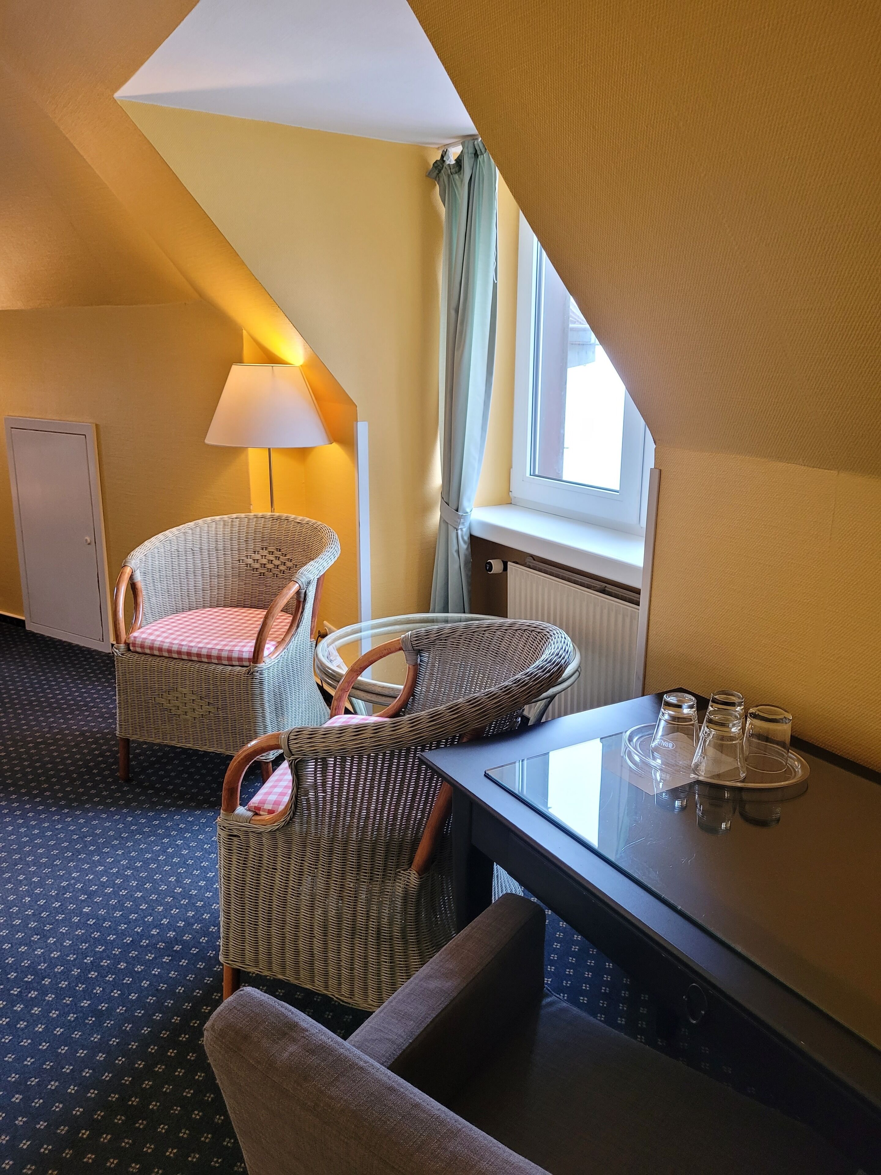 Habitación de hotel con dos sillas de mimbre, una mesa con vasos, una lámpara, una ventana con cortinas y un radiador.