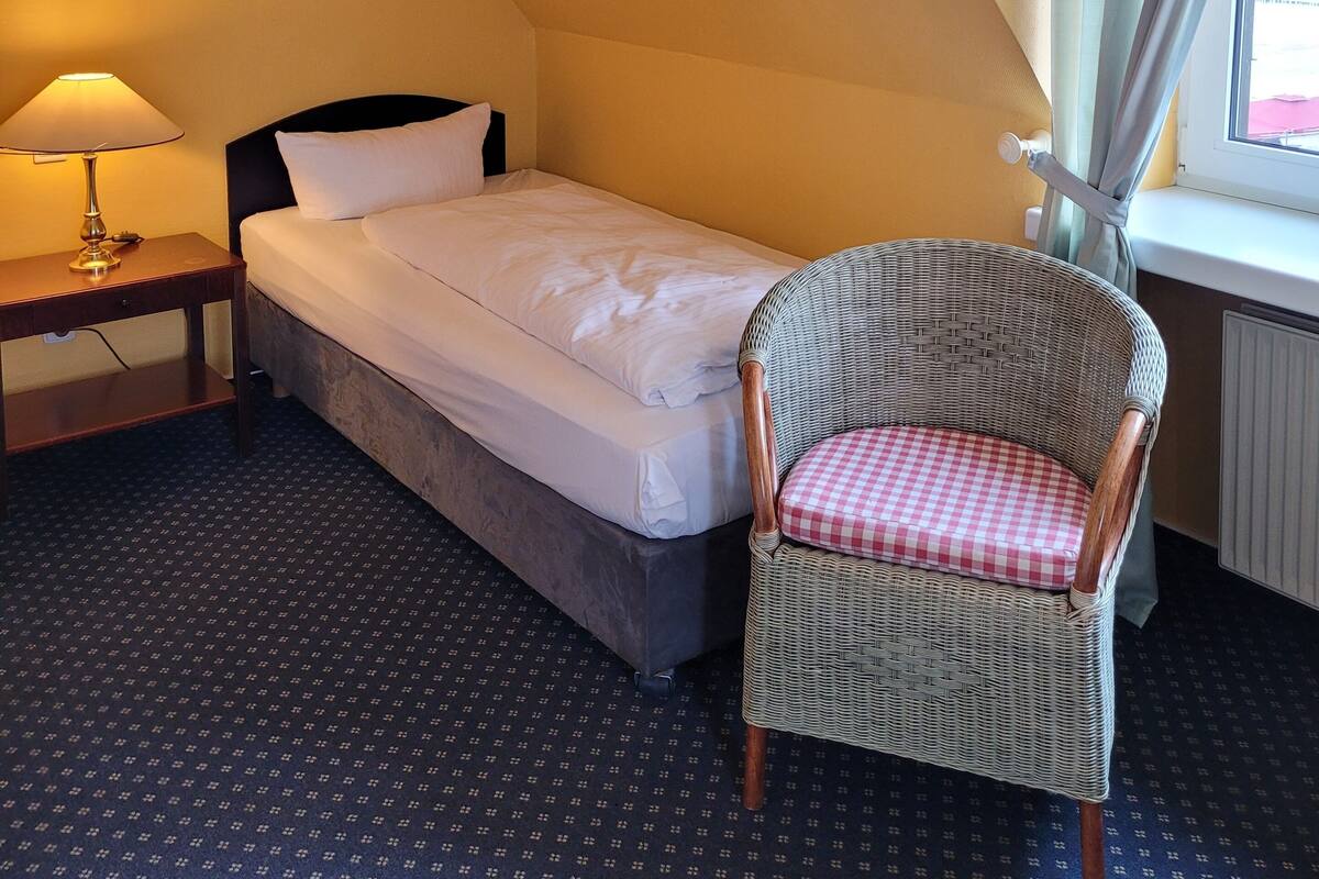 A small, single-bed room with a nightstand, a chair, a window with curtains, and a lamp.