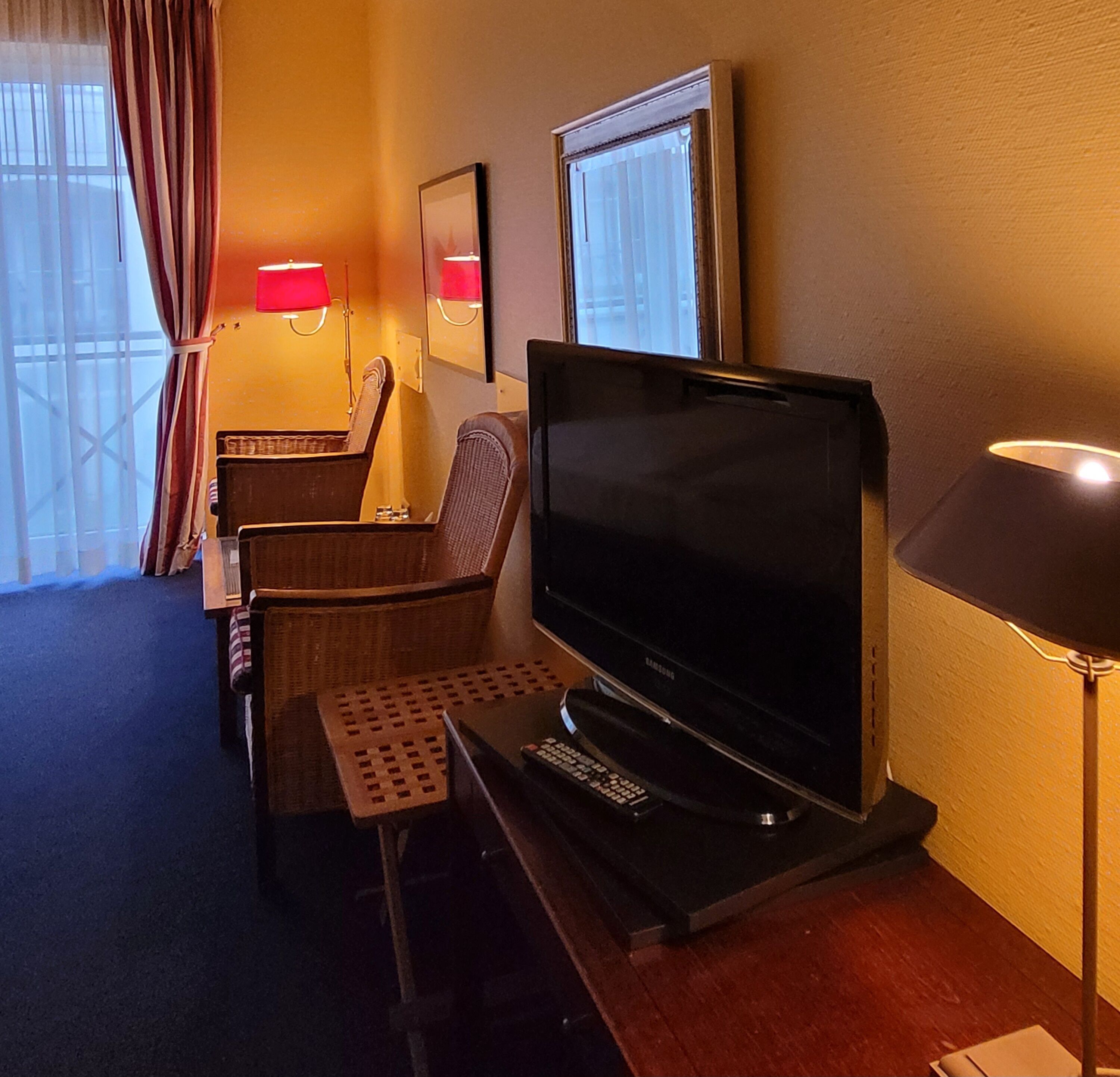 A hotel room with a flat-screen TV, a desk, a chair, a lamp, and a window with curtains.