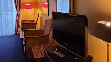 Ein Hotelzimmer mit Flachbildfernseher, Schreibtisch, Stuhl, Lampe und einem fenster mit Vorhängen.