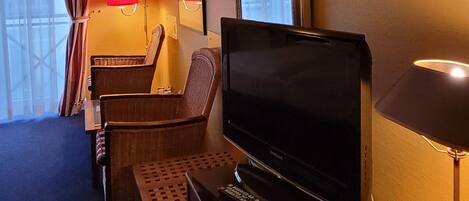 A hotel room with a flat-screen TV, a desk, a chair, a lamp, and a window with curtains.