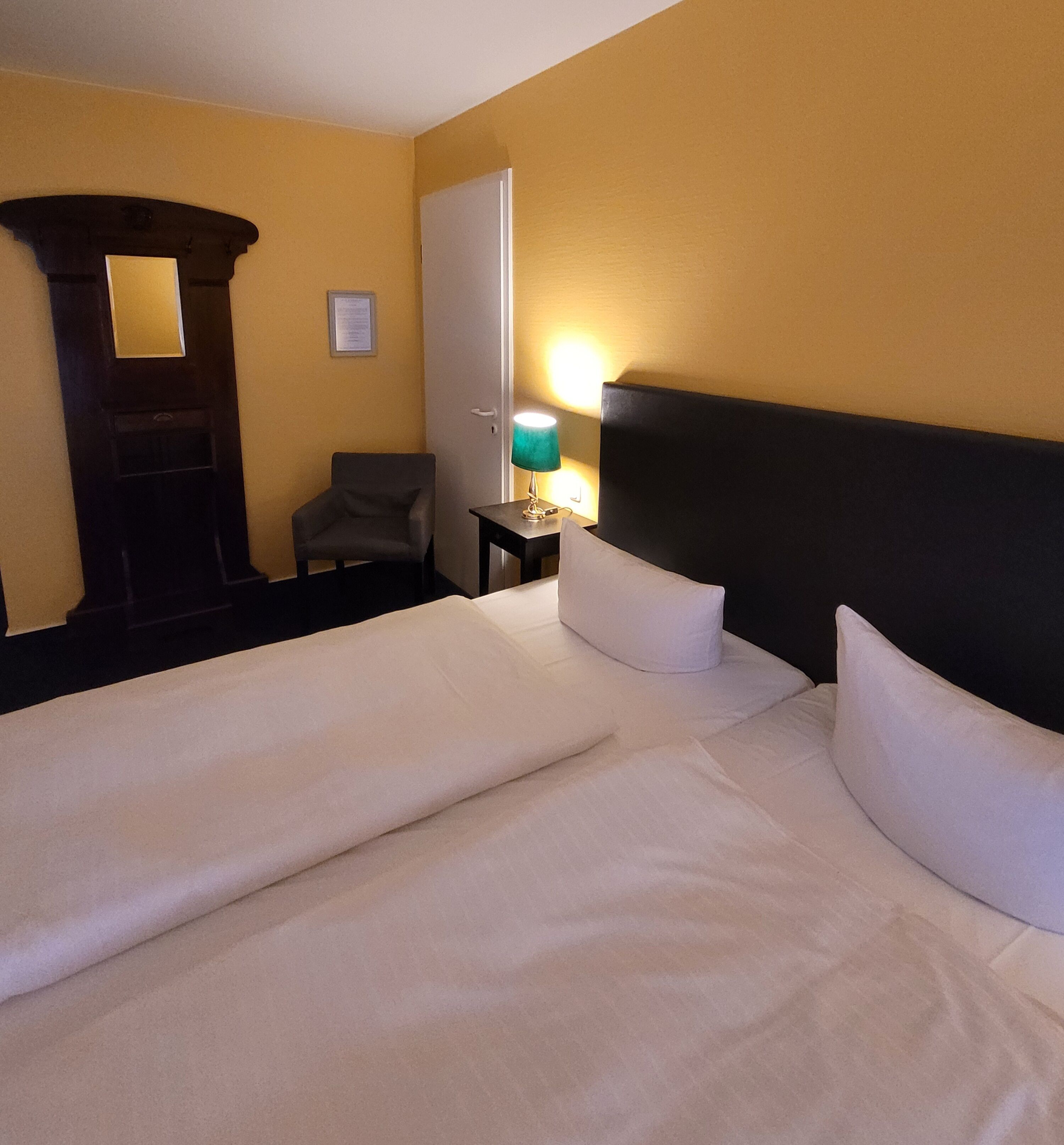 A hotel room with a large bed, a chair, a desk with a lamp, and a door.