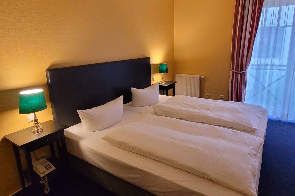 A hotel room with a large bed, two bedside tables, a green lamp, a telephone, and a window with curtains.