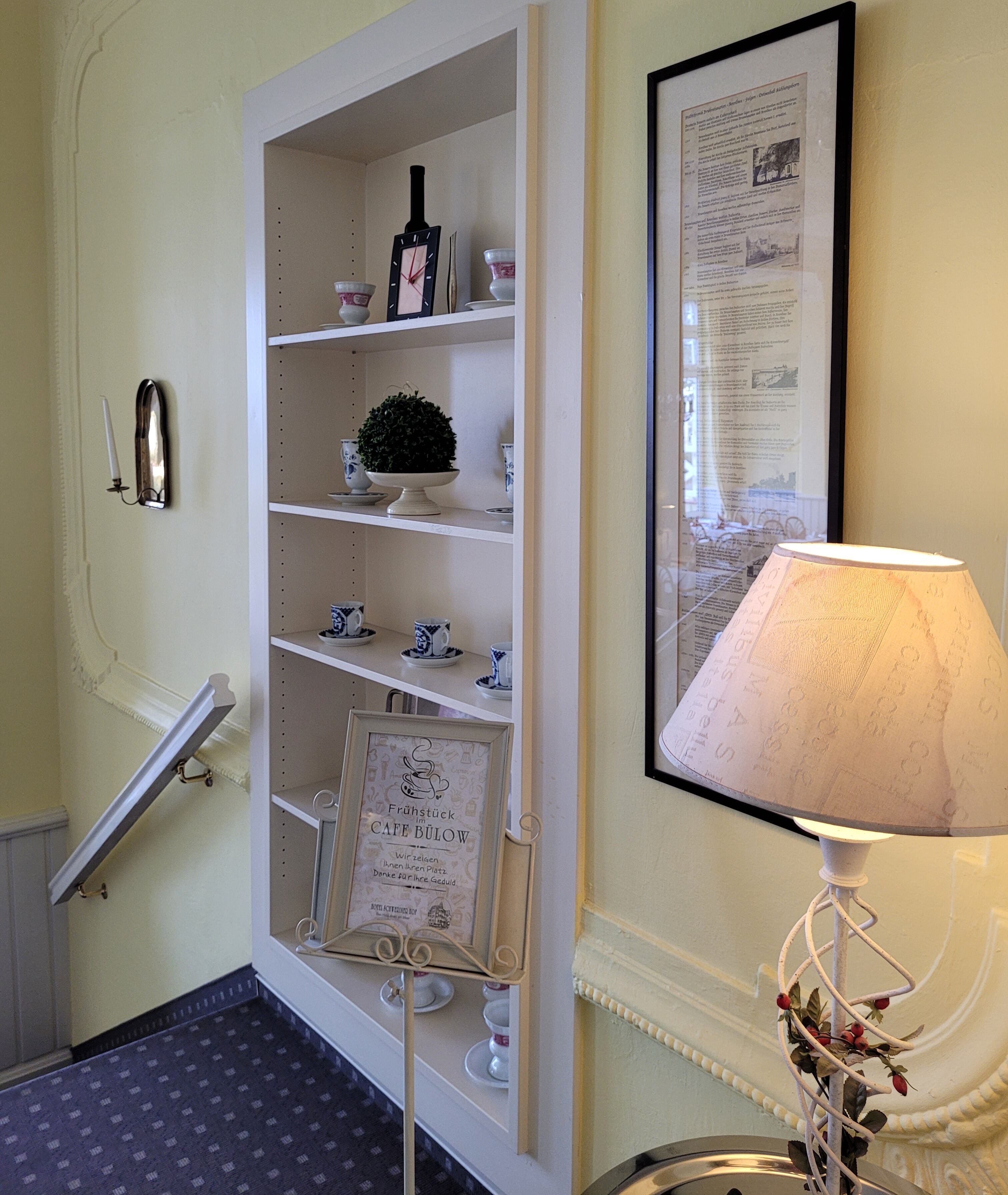 A hallway with a white shelf unit displaying tea cups, a framed menu, and a small potted plant.