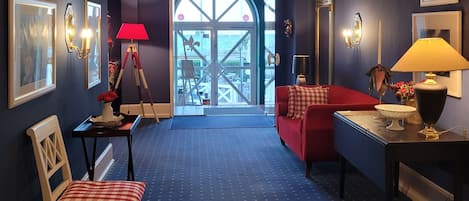 Una habitación con alfombra azul, sofá rojo, una silla de madera, una mesita con una lámpara y un espejo grande.