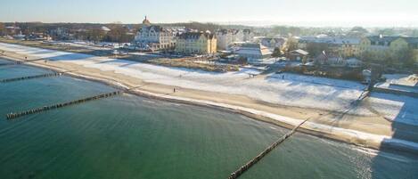 An aerial view of a coastal area with a sandy beach, a pier, and buildings in the background.