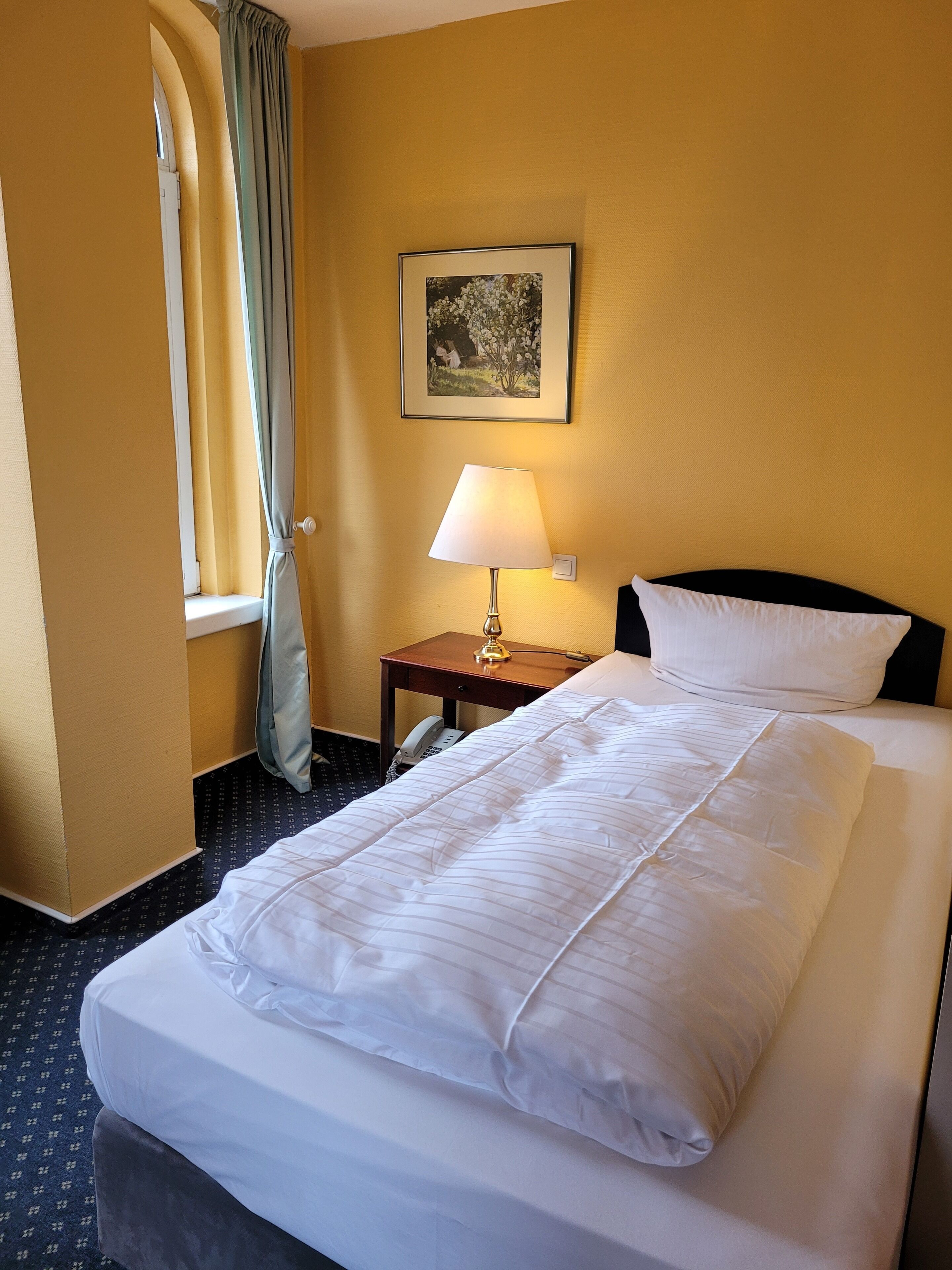 Een hotelkamer met een bed, een nachtkastje met een lamp, een telefoon en een ingelijst schilderij aan de muur.