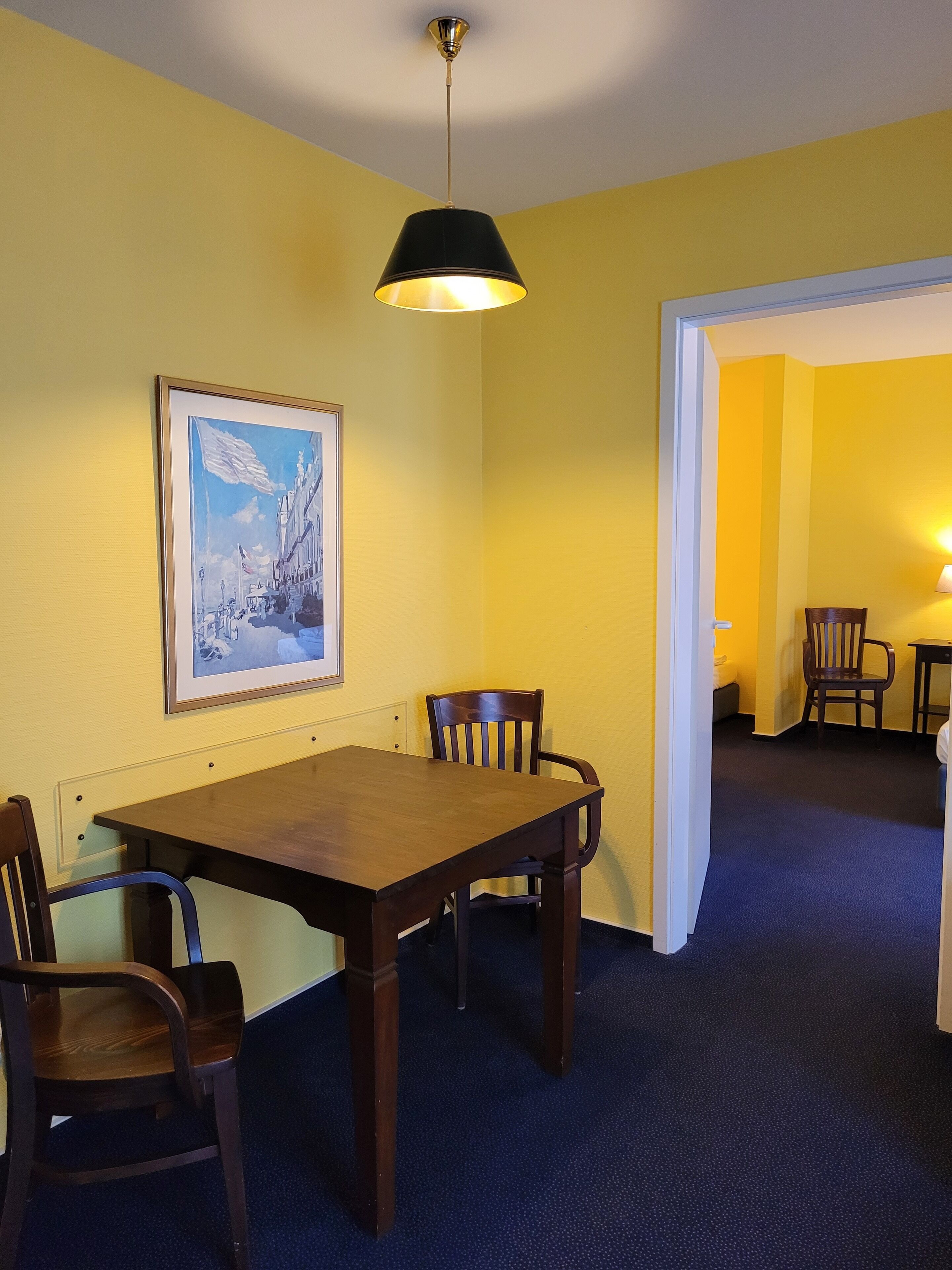 A dining area with a wooden table and chairs, a framed painting on the wall, and a ceiling light fixture.