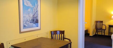 A dining area with a wooden table and chairs, a framed painting on the wall, and a ceiling light fixture.