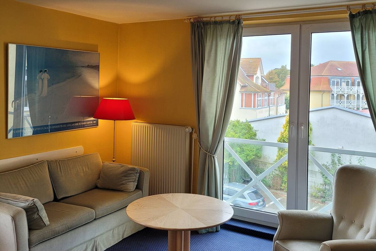 A hotel room with a sofa, a round table, a lamp, and a view of the street through large windows.