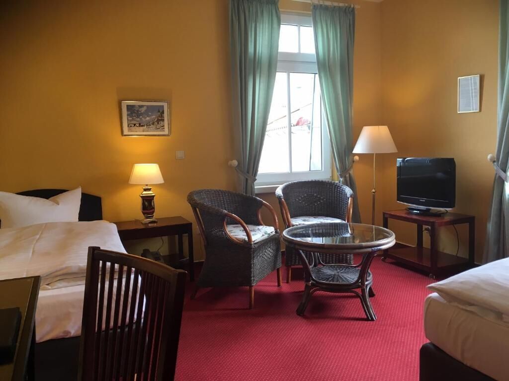 A hotel room with a bed, two wicker chairs, a small table, a TV, and a window with curtains.