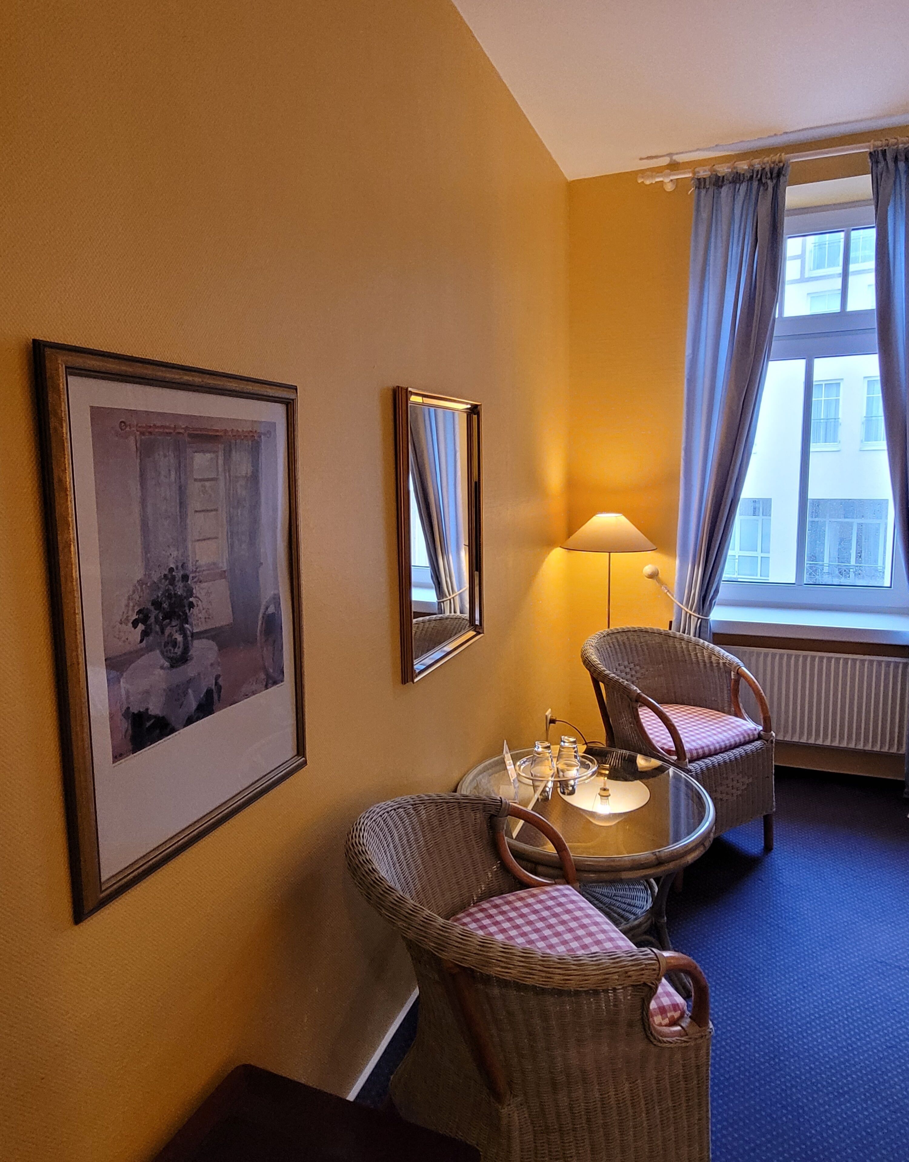 A hotel room with a bed, a window with curtains, a lamp, a chair, a table with a tray, and two framed pictures on the wall.