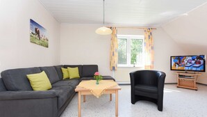 A living room with a grey sofa, a wooden coffee table with a vase, a black armchair, a television, and a window with curtains.