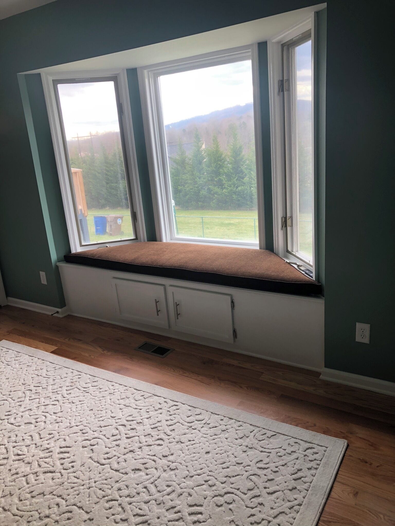 A room with a built-in bench under a large window, a white patterned rug, and a white cabinet.