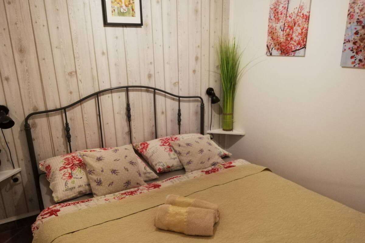 A bedroom with a wooden wall, a metal bed frame, floral bedding, a bedside lamp, and a vase with green plants.