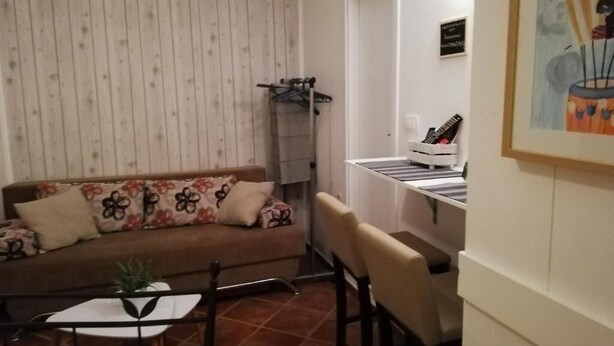 A cozy living room with a sofa, a small table, and a chair. There is a radiator, a painting on the wall, and a door leading to another room.