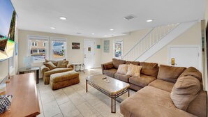 A spacious living room with a large sofa, ottoman, and coffee table. There is a television on the left and a staircase leading to another level. - ICONIC SEASIDE HEIGHTS BEACH HOUSE (Seaside Heights)