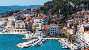 Un pueblo costero con un puerto deportivo, edificios de techos rojos y un paisaje montañoso al fondo.