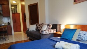 Un dormitorio con cama, mesita de noche, lámpara, sofá y una cocina al fondo.