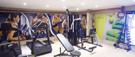 Academia com equipamentos de cardio, máquinas de musculação e um painel de espelhos com cartazes de ginástica.