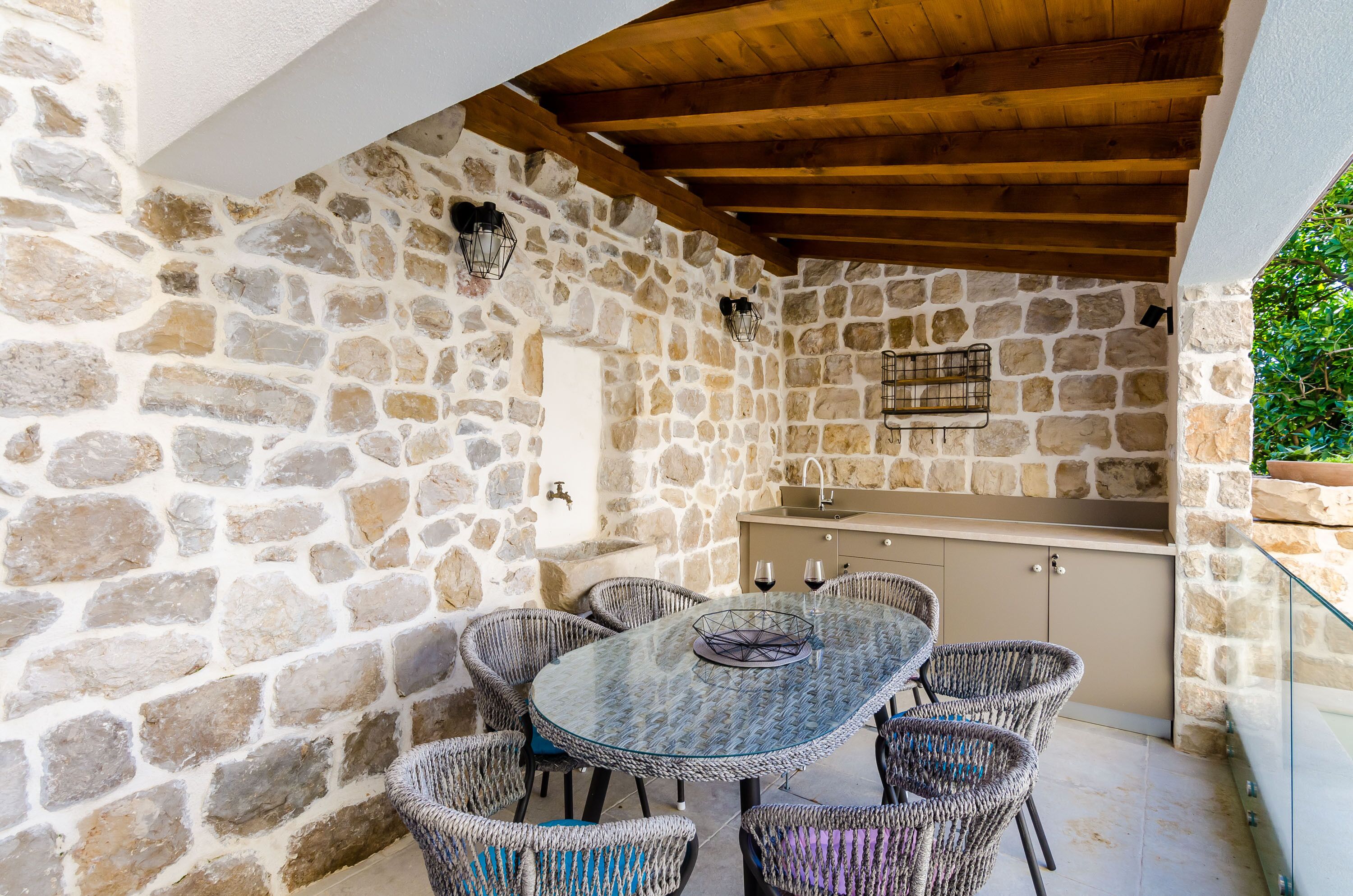 A stone-walled outdoor dining area with a round table and wicker chairs, a wooden ceiling, and a stone wall with a built-in sink.