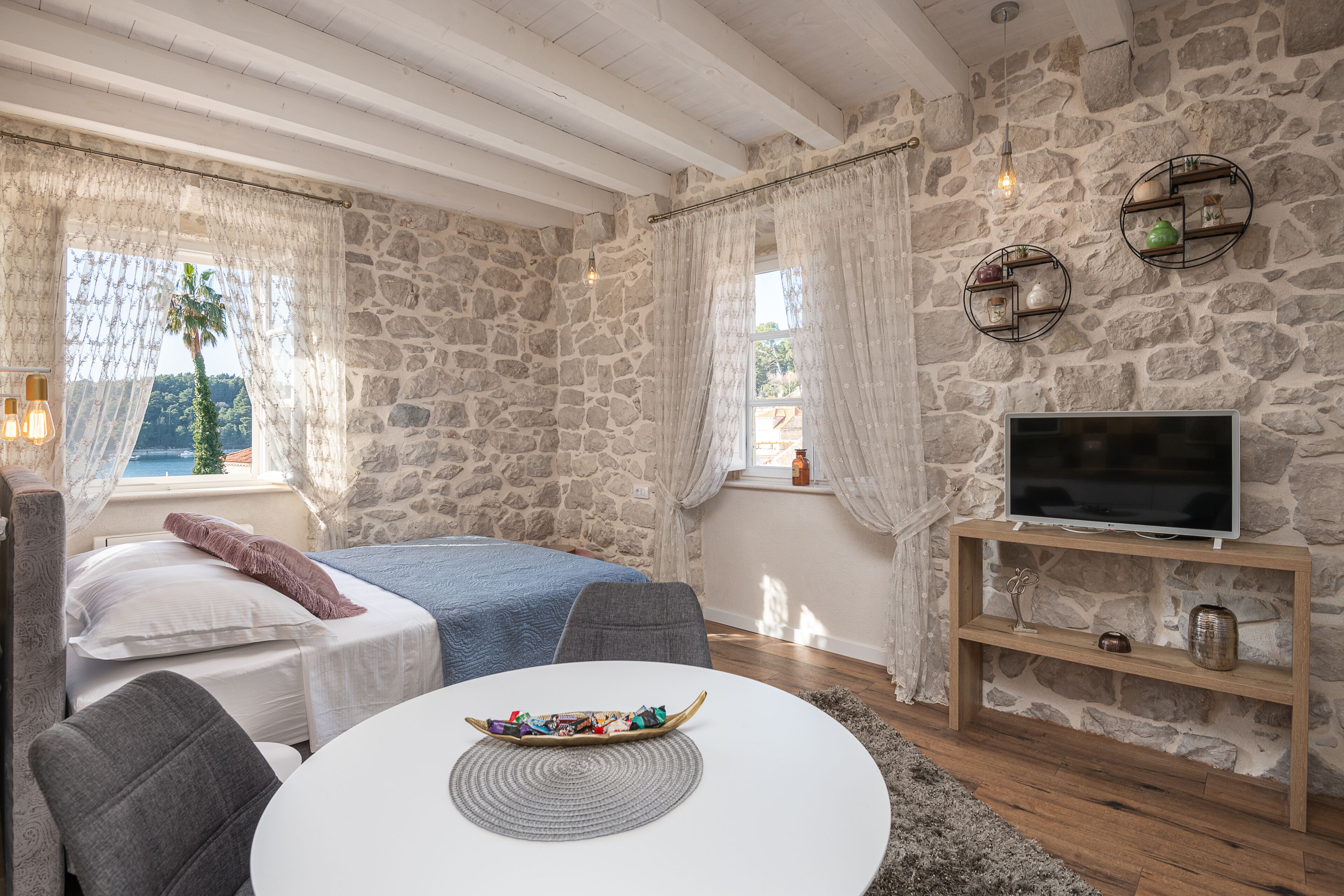 A cozy room with a stone wall, a bed with a blue blanket, a TV on a wooden stand, and a table with a bowl of snacks.