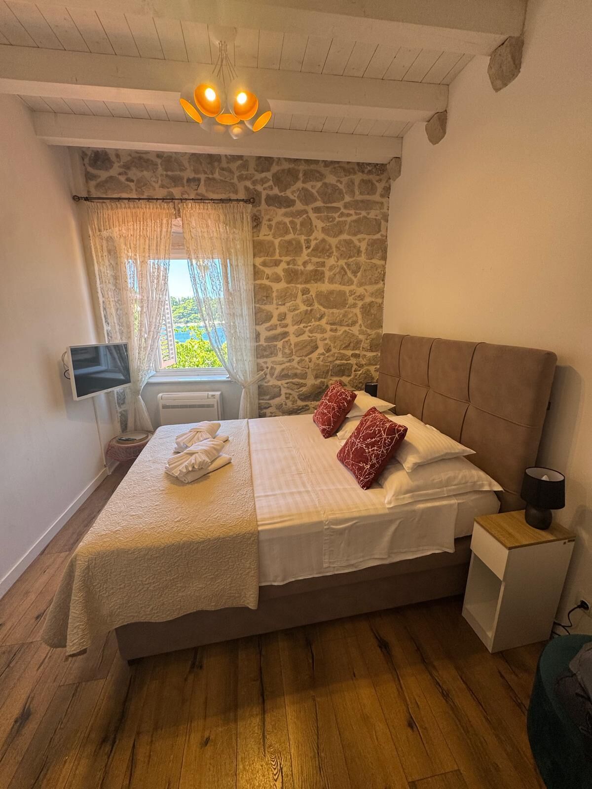 A bedroom with a stone wall, a bed with white bedding and red pillows, a wooden floor, a small bedside table with a lamp, and a window with curtains.