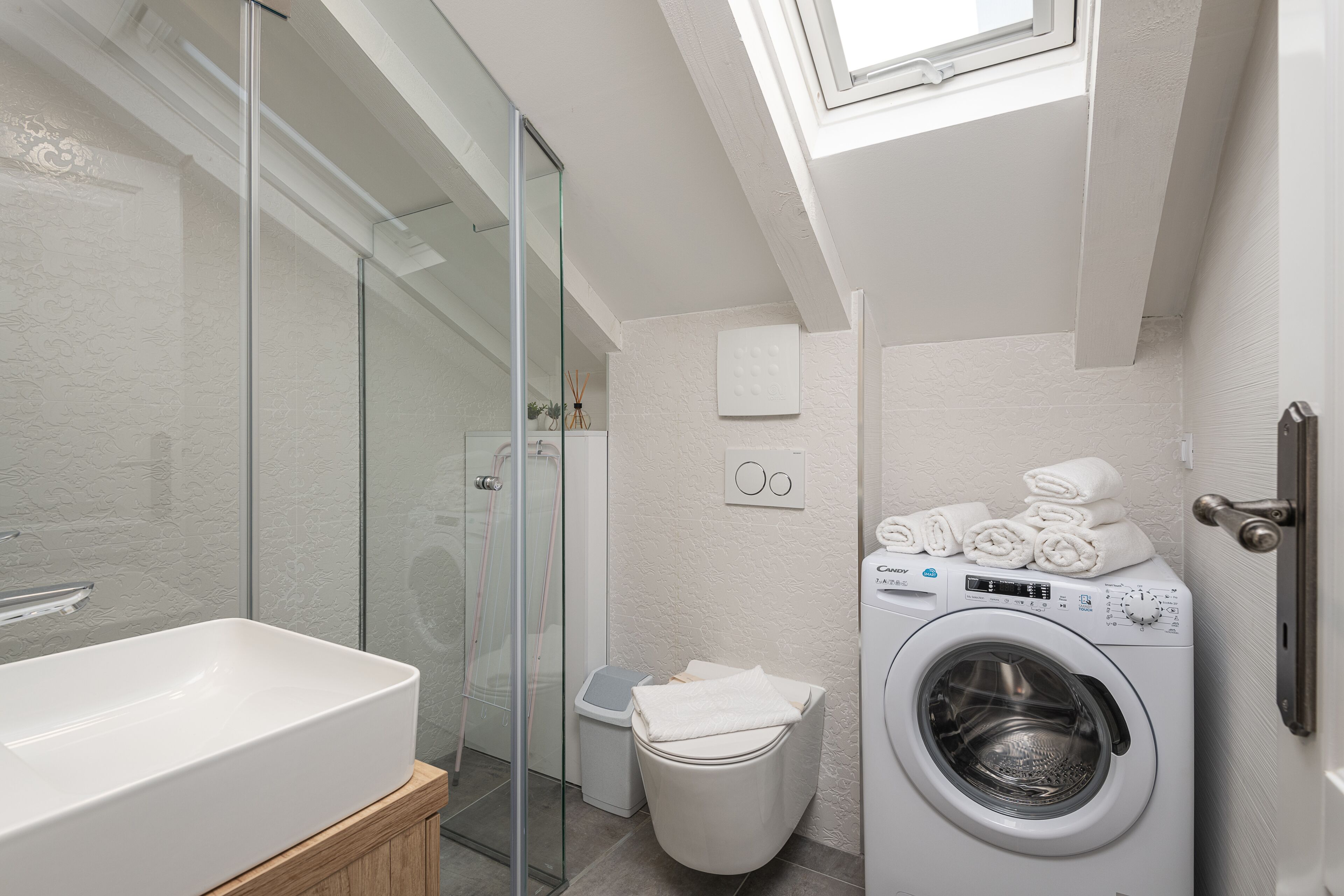 A modern bathroom with a glass shower enclosure, a white sink, a washing machine, and a small trash bin.