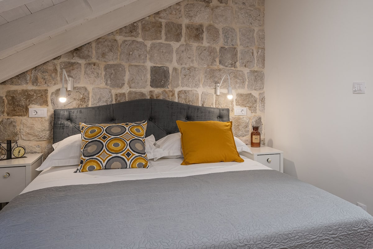 Ein Schlafzimmer mit Steinwand, grauem Polsterkopfteil, weißer Bettdecke und zwei Nachttischen mit Lampen.
