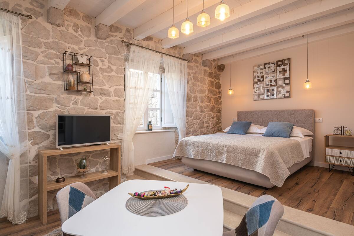 Ein gemütliches Schlafzimmer mit einer Steinwand, einem Bett mit weißer und grauer Bettdecke, einem Nachttisch mit Lampe und einem Fernseher auf einem Holzständer.