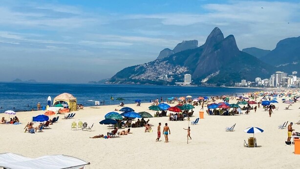 Ein überfüllter Strand mit Menschen, Sonnenschirmen und einem markanten Berg im Hintergrund.