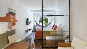 Ein modernes, helles Zimmer mit einem Holzesstisch, einer Couch, einer Hängemattenliege und einem großen Fenster, das viel Tageslicht hereinlässt.