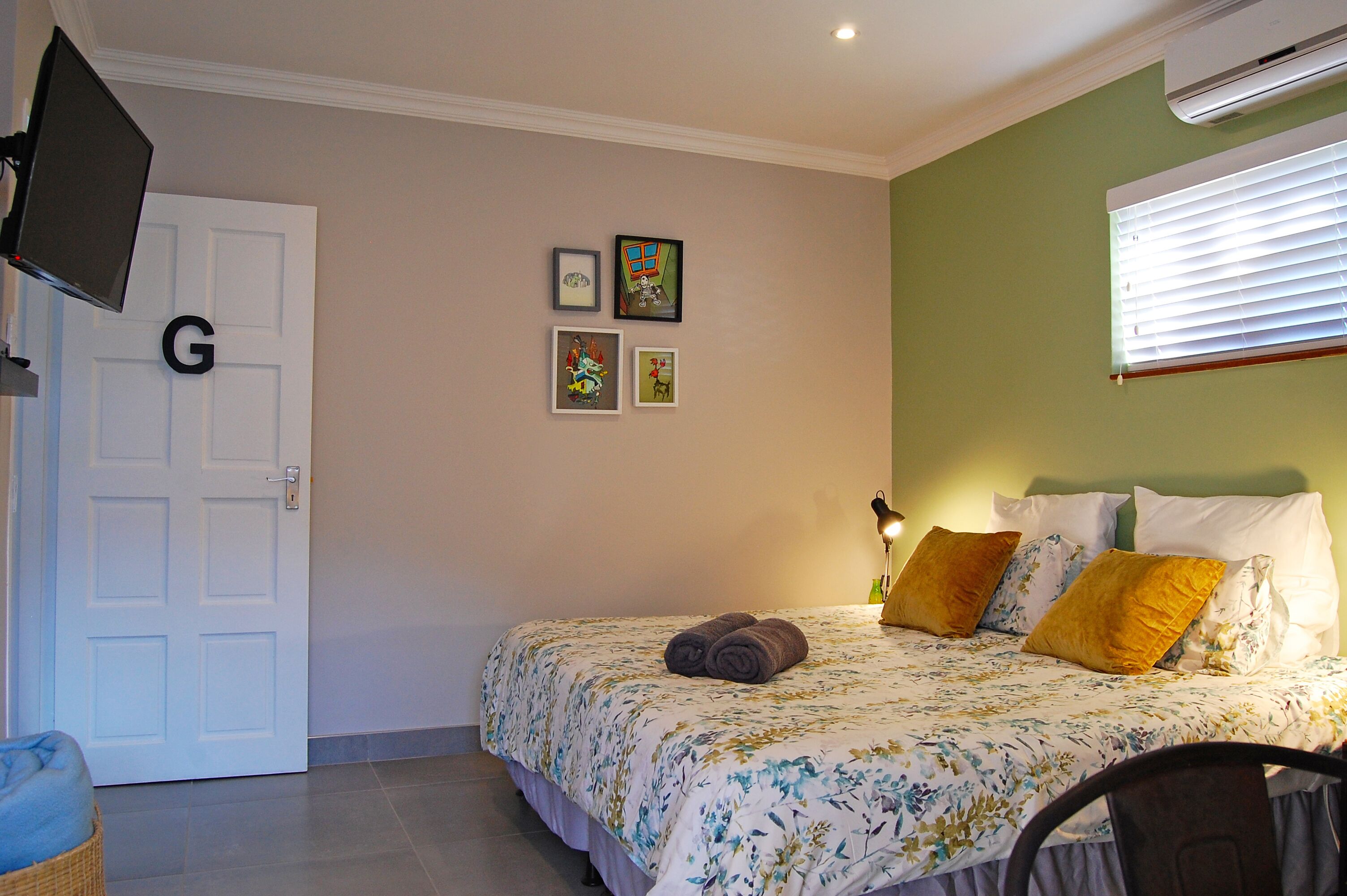A bedroom with a bed, a television mounted on the wall, and a door with a letter 'G'.