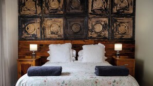 A bed with floral bedding, two white pillows, and two dark blue towels. There are two bedside lamps on wooden nightstands. The wall behind the bed features a distressed wood panel design. - Stonehaven Guest House (Clarens)