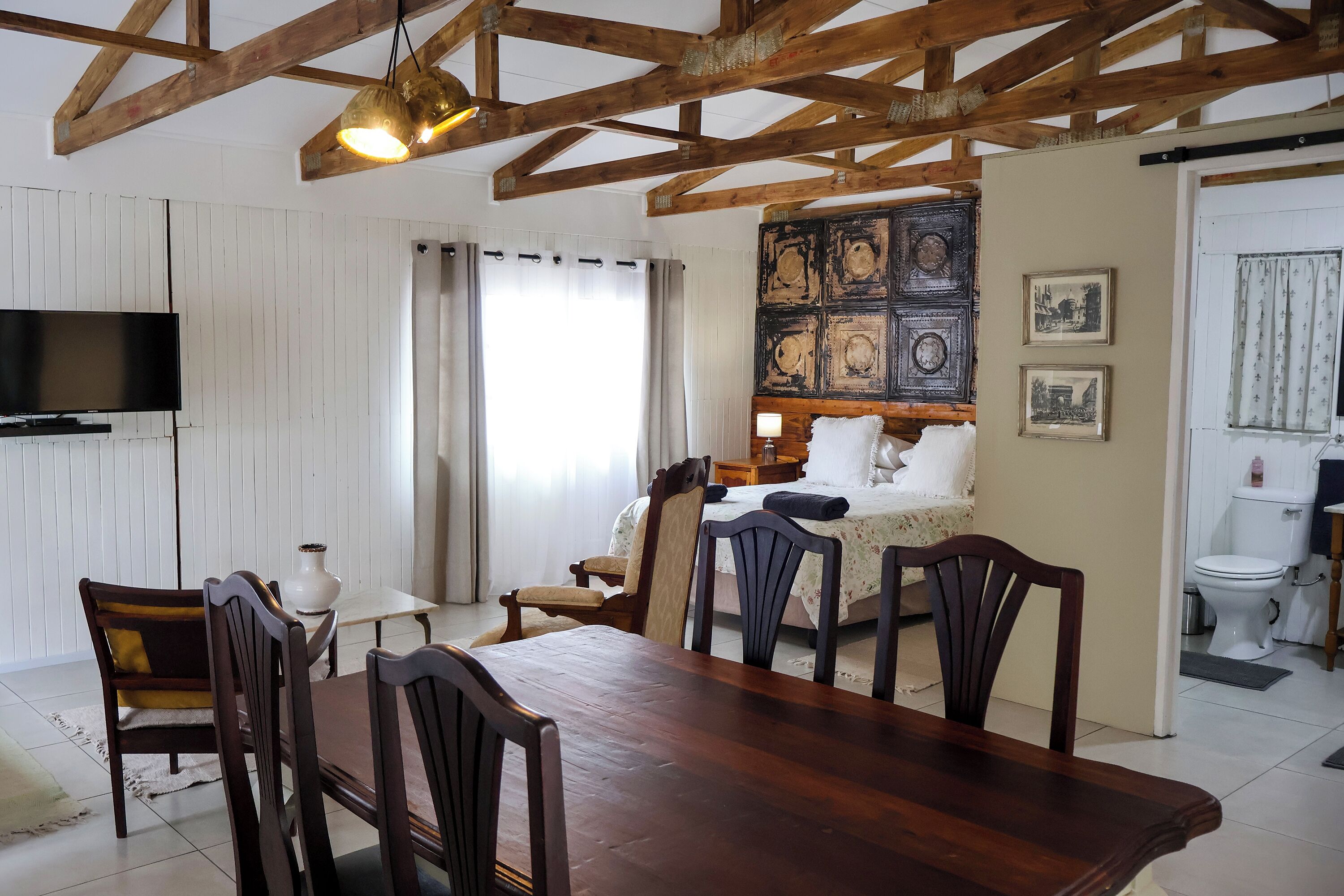 A room with a wooden ceiling, a dining table with chairs, a TV mounted on the wall, a bed with a nightstand, and a bathroom visible through an open door.