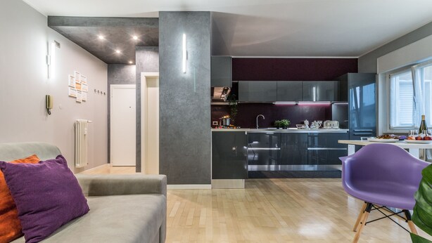 A modern kitchen with stainless steel appliances, a dining area with a purple chair, and a grey sofa with a purple pillow. - Family Apartments Verdi (Trento)