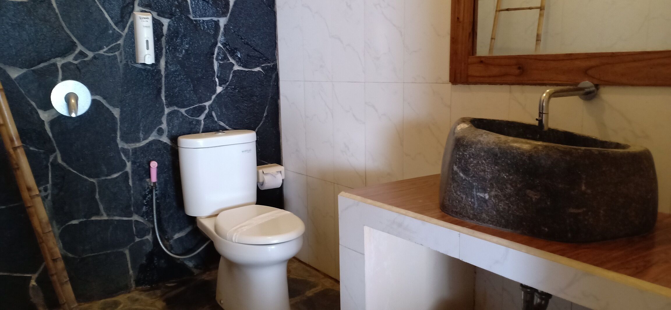 Ett badrum med en vägg av sten, en vit toalett, ett handfat med en skål av sten och en bänkskiva i trä.