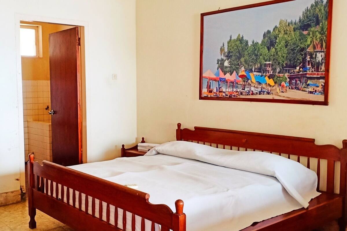A bedroom with a wooden bed, a door leading to a bathroom, and a painting of a beach scene on the wall.