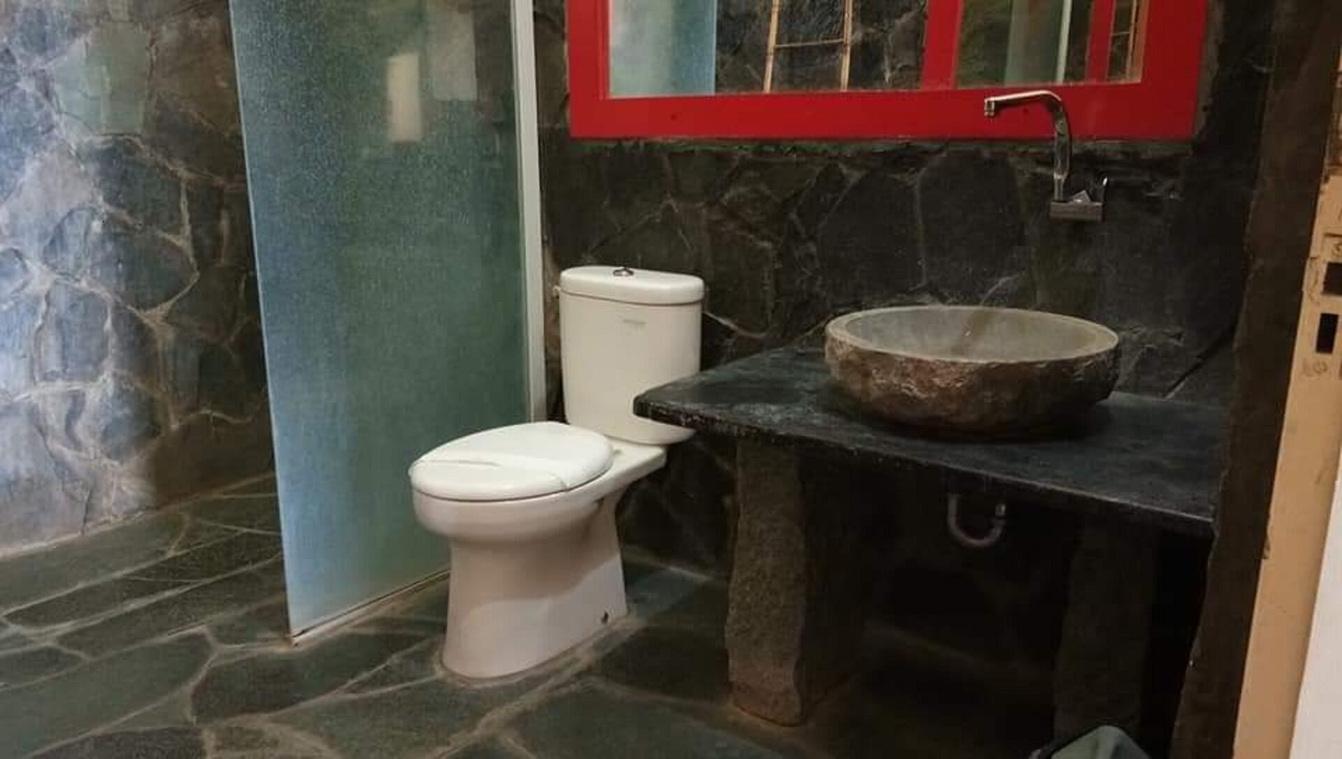 A bathroom with a white toilet, a stone sink, and a red-framed mirror.