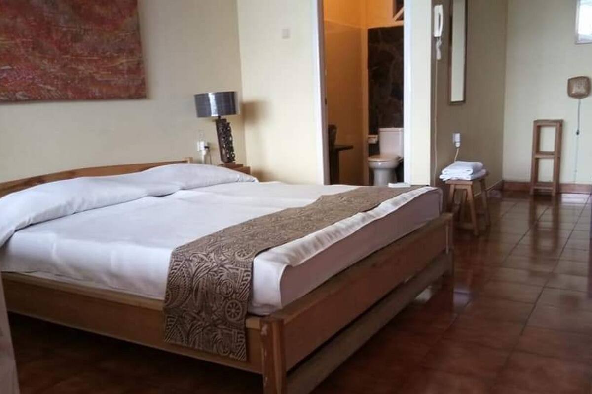 A hotel room with a large bed, a bedside table with a lamp, a small stool, and a desk with a chair.