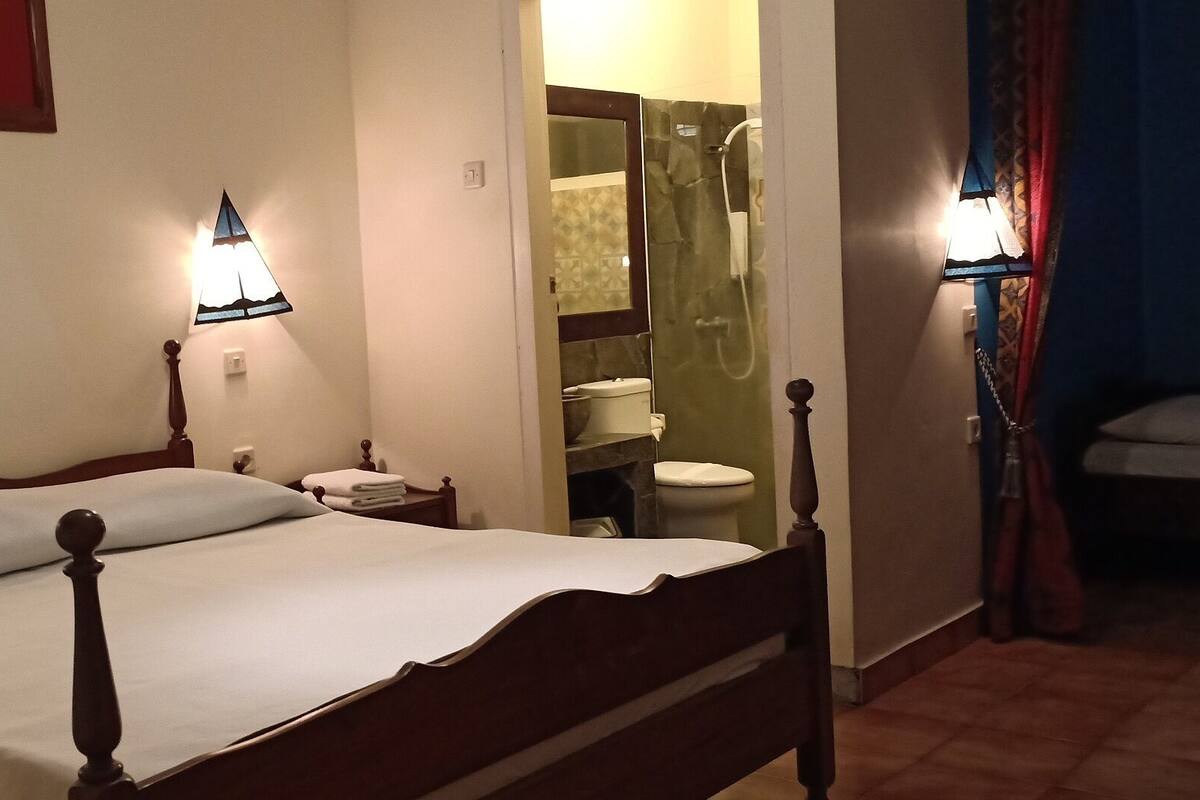 A bedroom with a wooden bed, two bedside lamps, a nightstand, and a bathroom visible through an open door.