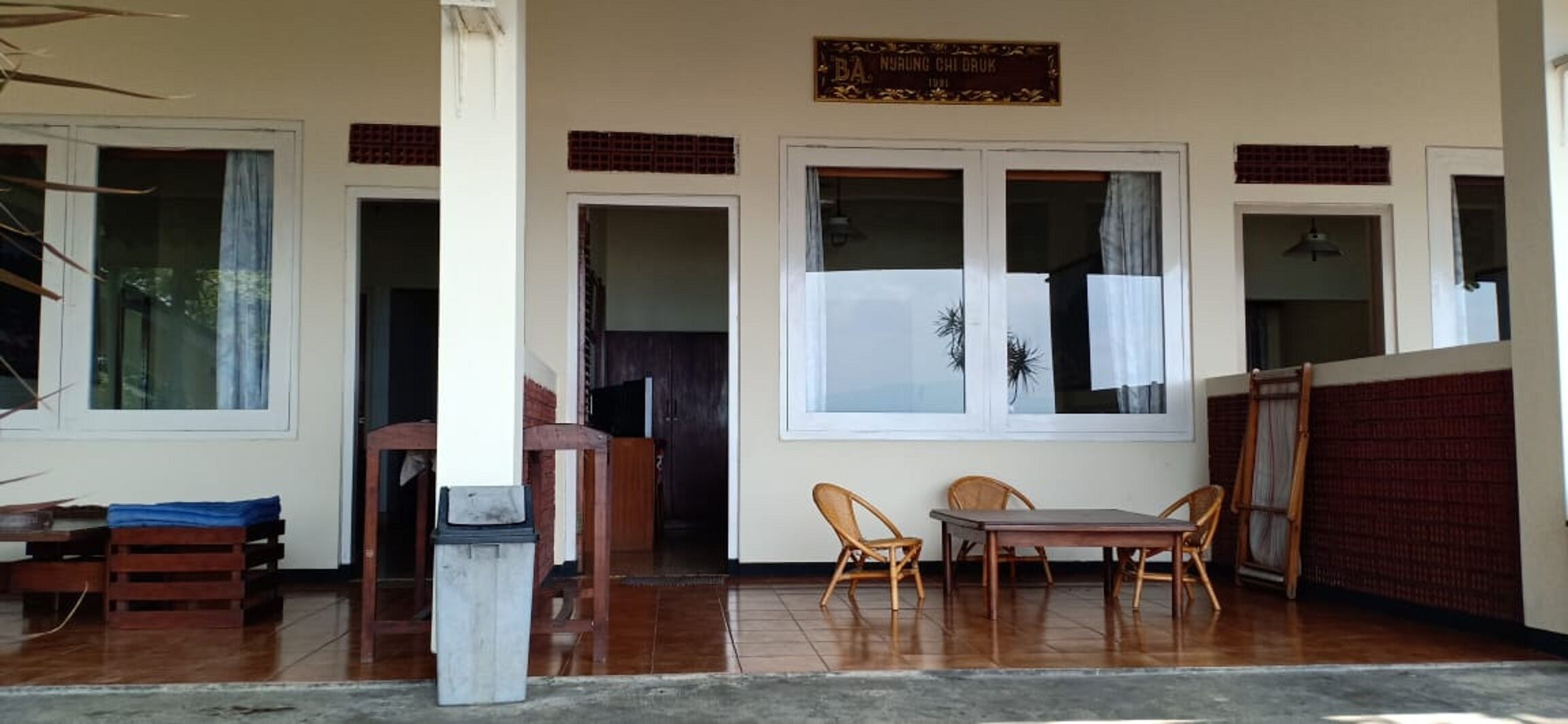 Una habitación con una mesa y sillas de madera, un cesto de basura y una ventana con cortinas.
